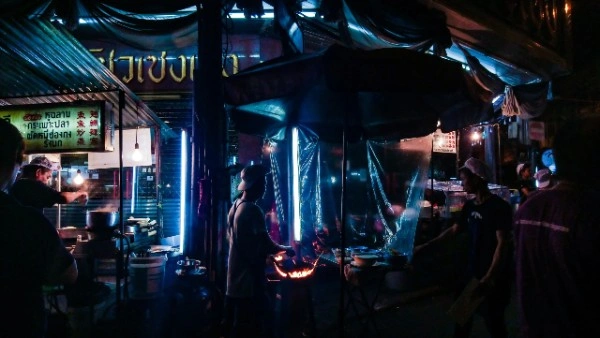 Atmospheric night street food market scene with vendors cooking under blue tarps and warm lighting, people gathering around food stalls