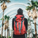 Traveler with red backpack walking through modern urban architecture with palm trees, representing someone ready for adventure through comprehensive preparation