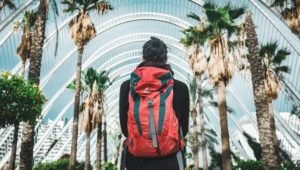 Traveler with red backpack walking through modern urban architecture with palm trees, representing someone ready for adventure through comprehensive preparation
