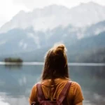 Woman with leather backpack standing peacefully at mountain lake shore, gazing at dramatic Alpine peaks reflected in calm water, representing the freedom and fulfillment of thoughtful budget travel