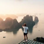 Person standing on wooden platform overlooking dramatic limestone karsts and emerald waters at golden hour, hand shading eyes while gazing into the distance, representing self-discovery and purposeful travel planning