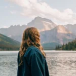 Person with flowing hair in denim jacket standing peacefully by pristine mountain lake, gazing at dramatic peaks reflected in calm water, representing serenity and overcoming travel anxiety