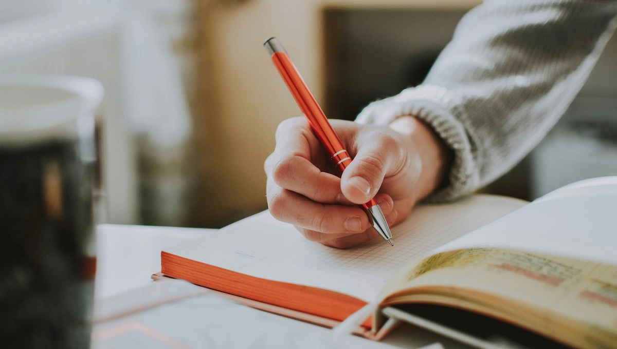 Person writing in an open journal with a red pen, capturing thoughts and reflections in a cozy indoor setting