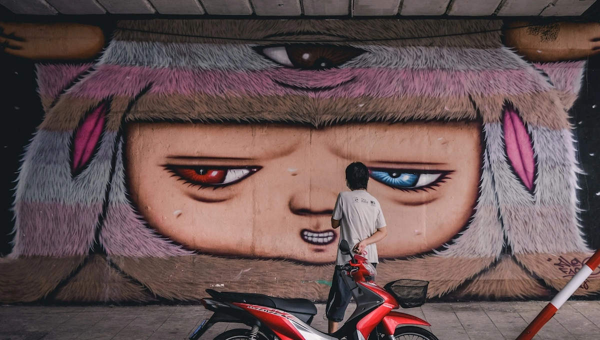 Person standing before large colorful street mural featuring an artistic character with mismatched eyes, discovering local creative expression